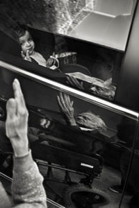 un niña dentro de un coche, en el reflejo del mismo se ve a una persona mayor saludando en silla de ruedas