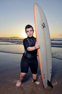 Imagen de un chico con acondroplasia en la playa con una tabla de surf